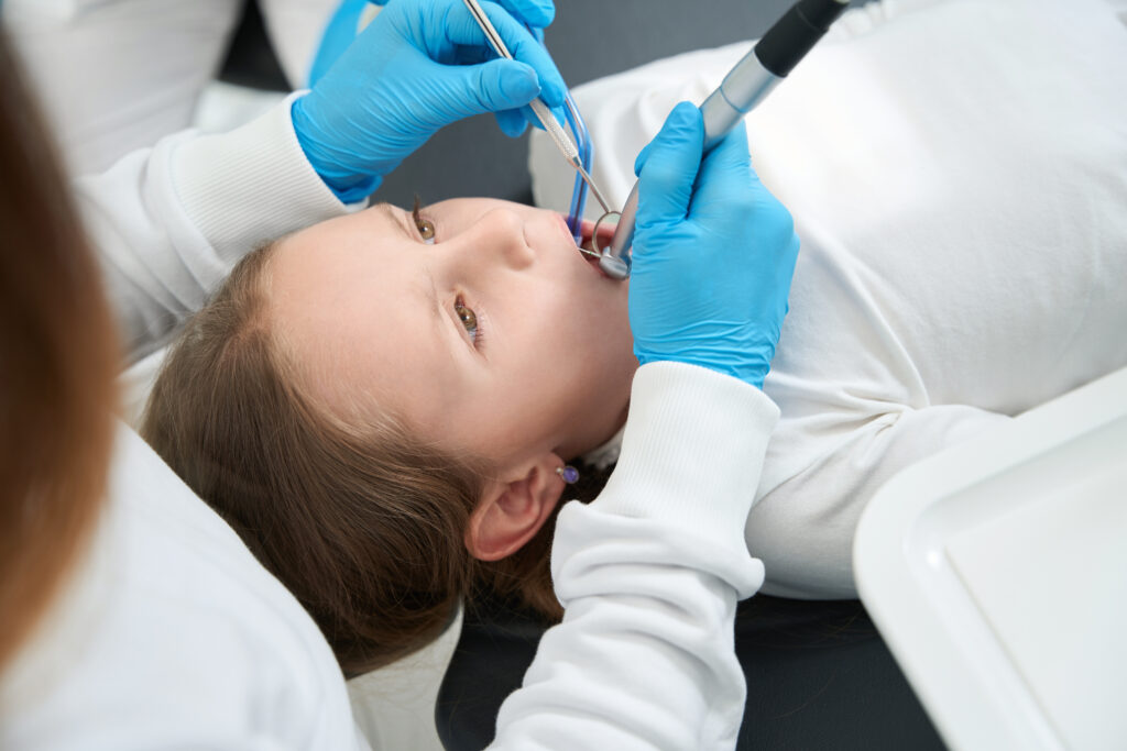 A child lies on a dental chair as a dentist, wearing blue gloves, examines their mouth with dental tools. The scene is calm and clinical.