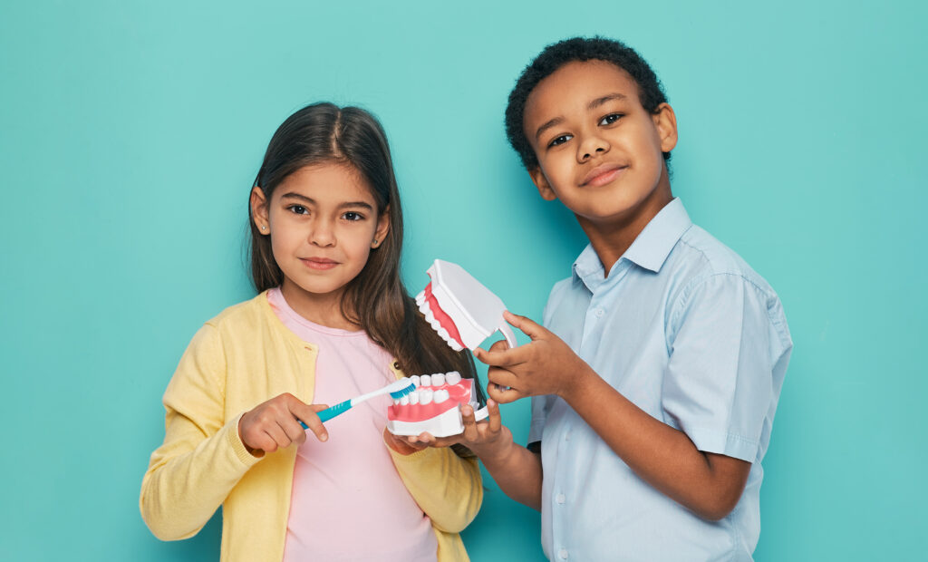 Two children stand against a teal background. The girl, in a yellow sweater, holds a toothbrush to a dental model. The boy, in a blue shirt, holds the model, smiling.